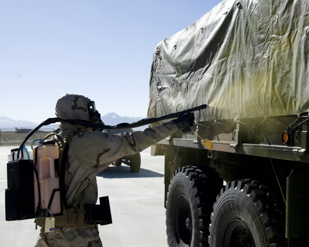 A soldier in a protective suit applies a spray to a military truck.