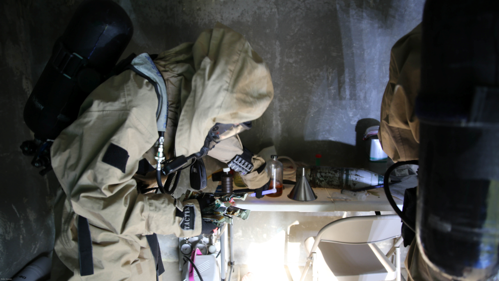 A U.S. Marine in chemical protective clothing checks places a purple chem light on a table with other equipment.