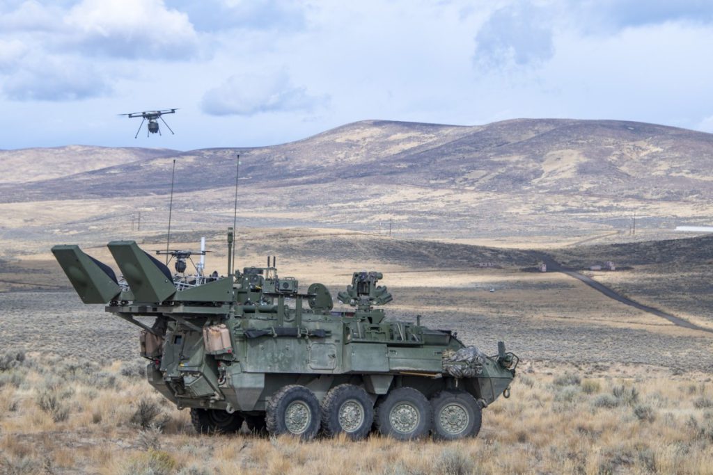 A military Stryker vehicle sits in a field. A drone flies overhead.