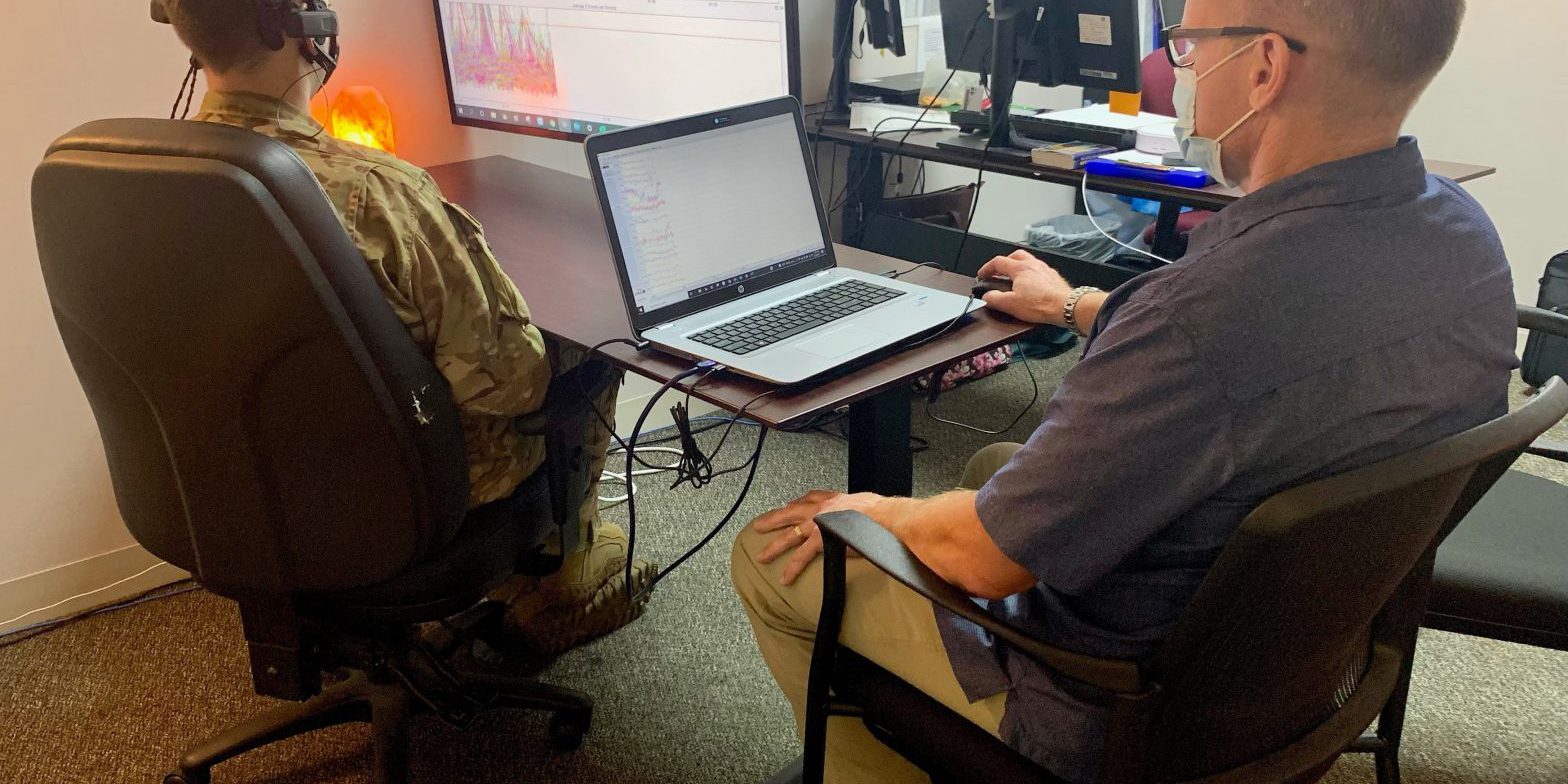 An Airman in uniform with sensors attached to his head sits at a computer screen. The test administrator, in civilian clothing, sits at a separate laptop.