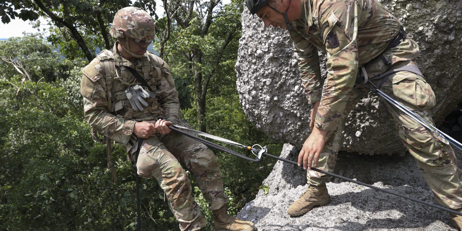 A Soldier prepares to rappel from a cliff. He is secured with a rope and leaning back getting ready to rappel down. Another Soldier looks on with his hand steadying the rope.