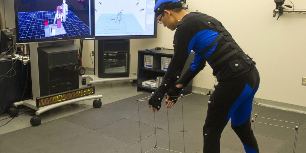 Image right: Marshall engineer Peter Ma, in the Marshall Virtual Environment Laboratory, wears a suit covered with spherical reflectors. This gear enables his body positions to be captured by a motion-tracking system, creating a model that is performing the same tasks in the virtual world that he is performing in the physical world. The human model in red on the screen represents the system-generated image of Ma's position, while the human model in light blue is a system-generated image of Kennedy Space Center engineer Jim Bolton, who is in Kennedy's Human Engineering Modeling and Performance laboratory. In this simulation, the two virtual humans picked up the models of the boxes shown on the floor and passed them back and forth. (NASA/MSFC/Emmett Given)