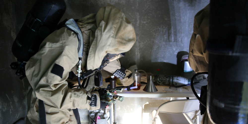 A U.S. Marine in chemical protective clothing checks places a purple chem light on a table with other equipment.